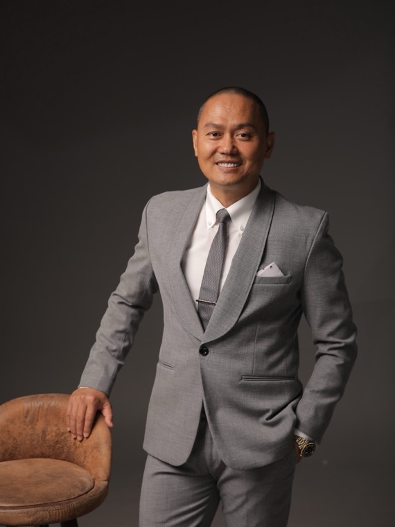 Joseph Duong seated in a chair in a light gray suit against a smoke-gray studio background, hands relaxed, looking slightly off camera.