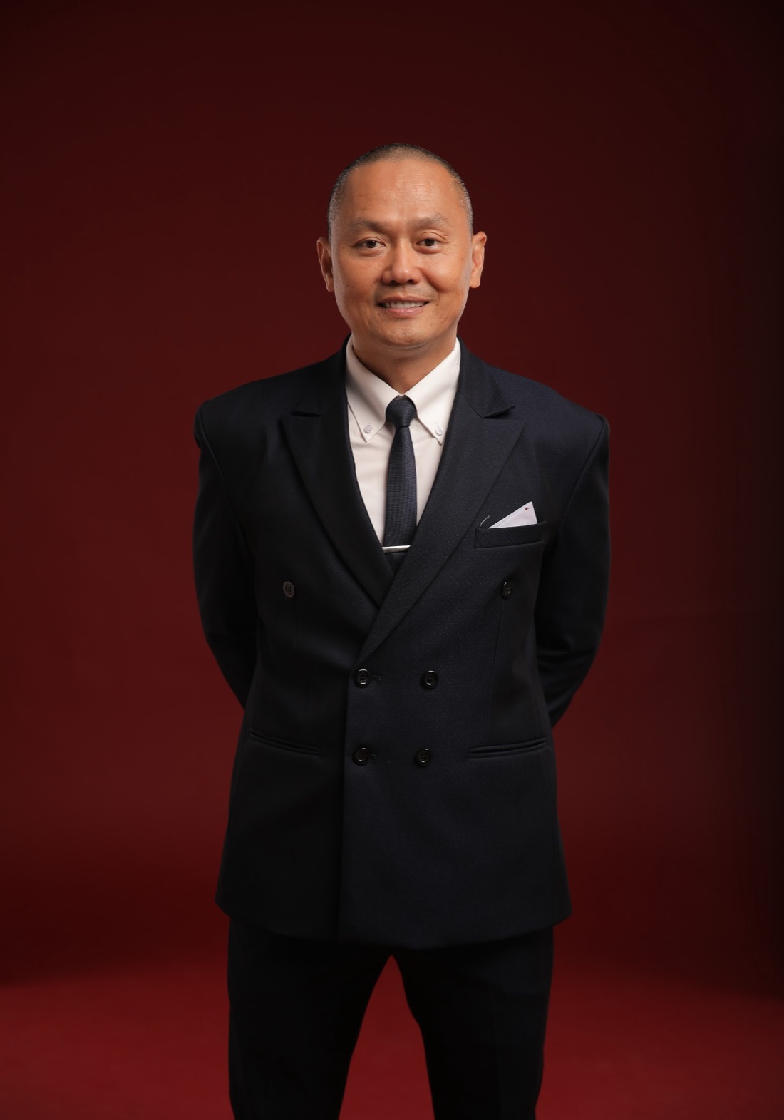 Joseph Duong, Orange County Realtor, standing with his arms crossed in a navy suit against a deep burgundy studio background, looking directly at the camera.
